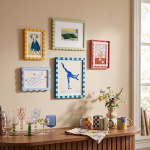 Könnte beinhalten: Eine Galerie-Wandgestaltung mit fünf bunten Bilderrahmen und verschiedenen Kunstdrucken, darunter ein Dalmatiner, eine blaue Figur und abstrakte Motive, über einem Holzkonsolentisch mit Gläsern und Tassen platziert.