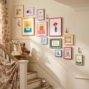 Op de afbeelding: Een galerijmuur met ingelijste kunstprints met kleurrijke illustraties van fruit, bloemen en desserts. De frames zijn in verschillende kleuren, waaronder geel, roze en groen. Een trap met een bloemengordijn en decoratieve hondenbeeldjes is ook zichtbaar.