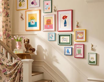 May include: A gallery wall featuring twelve colorful framed art prints with various illustrations including a cake, flowers, and a matcha latte. The frames are in assorted bright colors and are arranged on a light-colored wall above a staircase.