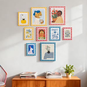 May include: A collection of framed art prints with various illustrations. The prints feature a cat with a beret, a person with flowers, a rabbit, lemons, fish, and the words "Cheers!" and "Self Care." The frames have colorful striped and checkered patterns.