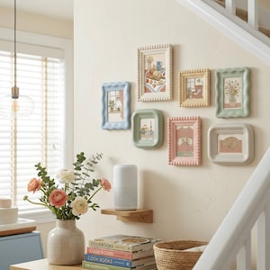 Peut inclure: Sept cadres décoratifs aux couleurs pastel sont accrochés sur un mur crème. Les cadres, aux bords festonnés, contiennent des illustrations de scènes domestiques. Un vase de fleurs et une pile de livres de cuisine sont au premier plan, créant une ambiance chaleureuse.