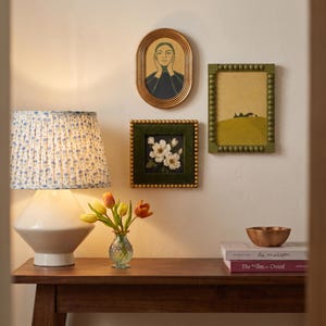 May include: A white table lamp with a blue floral pleated shade, a small glass vase with yellow and orange tulips, and three framed wall art pieces. A copper bowl sits on two stacked books titled la maison and The Inn Crowd on a wooden table.