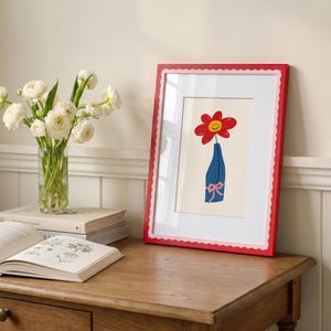 May include: A framed art print featuring a red flower with a yellow smiley face center in a blue vase with a pink bow. The frame is red with a scalloped edge and a white mat. The print is displayed on a wooden surface next to a glass vase of white flowers.