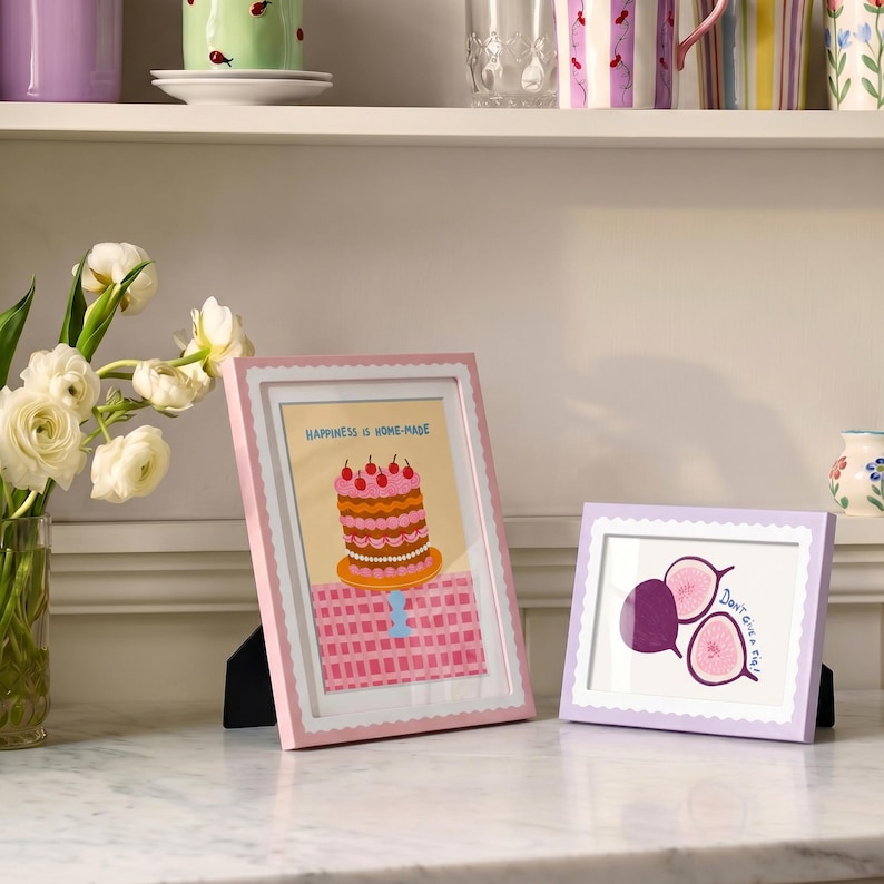 May include: Two rectangular picture frames with scalloped edges. The taller pink frame displays a cake illustration with the text "HAPPINESS IS HOME-MADE". The shorter lavender frame features a fig illustration with the text "Don't give a fig!".