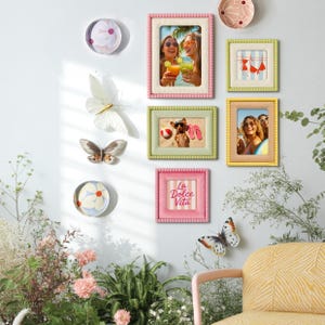 May include: A collection of decorative wall art, including framed photos, butterfly ornaments, and floral plates. The frames are in various colors, including pink, green, and yellow. One frame has the text "La Dolce Vita".