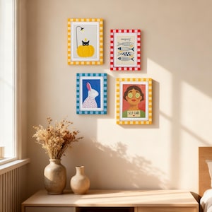 May include: Four framed art prints with colorful designs hang on a beige wall. The prints feature a black cat on a pumpkin, fish, a rabbit, and a woman with cucumber slices on her eyes. The frames have checkered patterns.