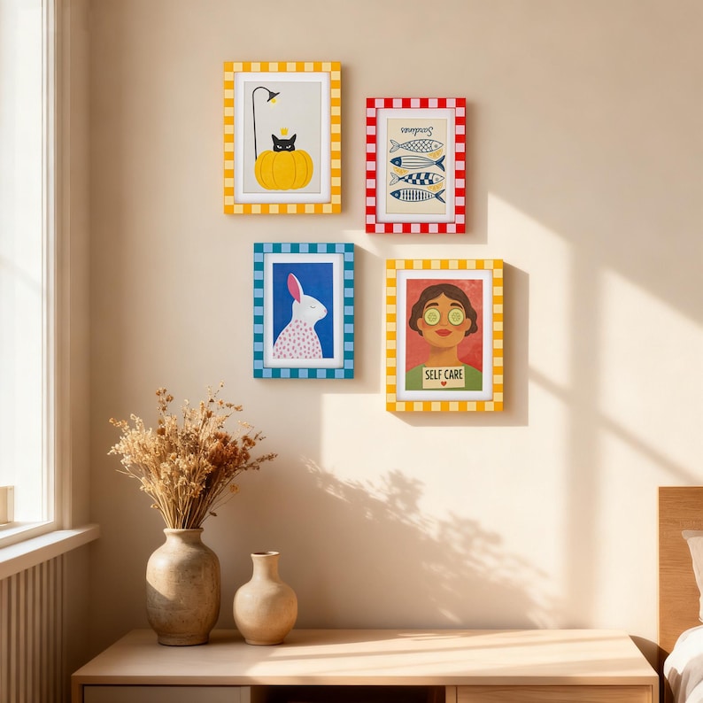 May include: Four framed art prints with colorful designs hang on a beige wall. The prints feature a black cat on a pumpkin, fish, a rabbit, and a woman with cucumber slices on her eyes. The frames have checkered patterns.