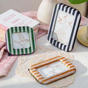 Könnte beinhalten: Drei dekorative Bilderrahmen mit Vogelillustrationen. Ein Rahmen hat grün-weiße Streifen, ein anderer marineblau-weiße Streifen und der dritte orange-weiße Streifen. Im Hintergrund ein Buch mit dem Text "THE WORLD is like a mirror".