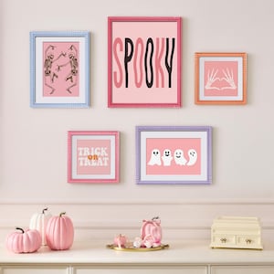 Op de afbeelding: Een verzameling ingelijste Halloween-kunstprints en decoratieve pompoenen. De prints tonen skeletten, geesten en de woorden "Spooky" en "Trick or Treat". De pompoenen zijn in verschillende roze en witte tinten.