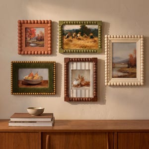 May include: A gallery wall arrangement featuring five rectangular framed artworks with beaded borders. The frames are in shades of terracotta, olive green, dark brown, and cream. The prints depict autumn landscapes, a bowl of pears, and pumpkins.
