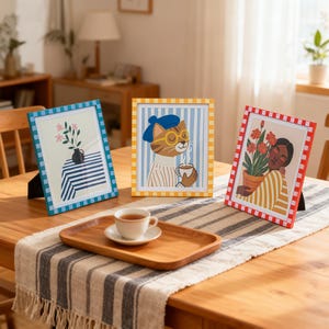 May include: Three framed art prints on a wooden table. The prints feature illustrations of a cat, flowers, and a person. The frames are blue, yellow, and red with checkered patterns. A wooden tray with a teacup and saucer sits on a striped table runner.