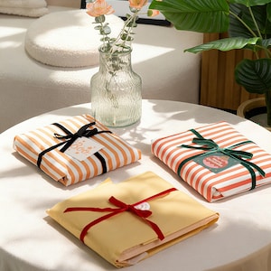 May include: Three wrapped gifts on a white table. Two are wrapped in orange and white striped paper with black and green ribbons. The third is wrapped in yellow paper with a red ribbon. A glass vase with flowers is in the background.