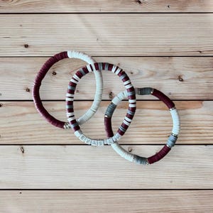May include: Three beaded bracelets in maroon, white, and gray. The bracelets are stacked on top of each other. The white beads have the words "TEXAS A&M" spelled out on them.