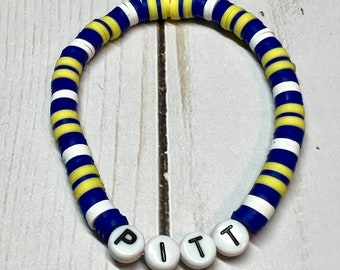 May include: A blue and yellow striped bracelet with white beads and the word "PITT" spelled out in white letters.