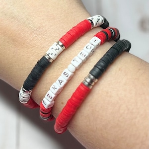 May include: Three bracelets stacked on a wrist. The top bracelet is black and white with white beads that spell "TECH". The middle bracelet is red and white with white beads that spell "TEXAS". The bottom bracelet is black and red with black beads.