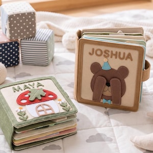 May include: Two personalized fabric books with felt appliques, one featuring a mushroom house labeled MAYA and one featuring a bear labeled JOSHUA. The set includes three patterned fabric cubes, two crocheted rattles, and a crescent moon plush toy.
