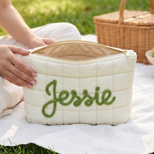 May include: Cream-colored quilted cosmetic bag with the name "Jessie" embroidered in green script. The bag has a gold zipper and is open, revealing the interior. A picnic basket and fruit are in the background.
