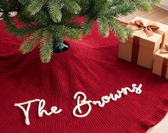 May include: A circular, deep red knitted Christmas tree skirt featuring the white embroidered text "The Browns" in a cursive script. The skirt is positioned under a green pine tree, accompanied by a small snowman figurine and gift boxes.