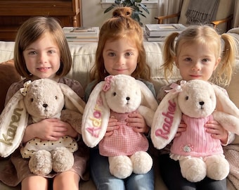 May include: Three plush stuffed bunnies with long ears, each featuring embroidered names on the ear. The first bunny is beige with a polka dot dress, the second is white with a pink gingham dress, and the third is white with a pink sweater.