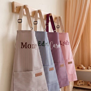 Puede incluir: Cuatro delantales a rayas colgados en un perchero de madera. Los delantales son marrón, azul, morado oscuro y rosa claro, cada uno con un bolsillo frontal con una pequeña etiqueta marrón que dice apron. Los nombres Monty, Ed, Carol y Lydia están bordados.