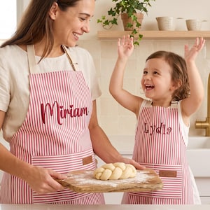 Op de afbeelding: Twee bijpassende schorten met een rood-wit verticaal gestreept patroon. Elk schort heeft een voorzak met een klein bruin label met de tekst "apron" en is geborduurd met een naam in rode schrijfletters: "Miriam" en "Lydia".