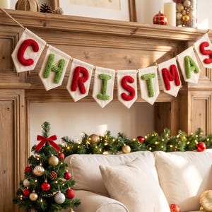 May include: A decorative banner featuring nine off-white fabric pennants strung together, each displaying a single letter in raised, textured knit yarn. The letters spell out the word CHRISTMAS in alternating red and green colors.