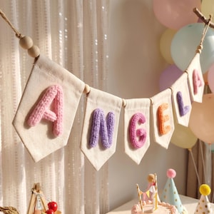 May include: A decorative banner featuring seven off-white canvas pennants strung together with wooden beads. Each pennant displays a single, chunky, hand-knitted letter in pastel colors, spelling out the name ANGELA in pink, purple, and orange.