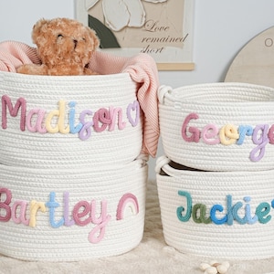 May include: Four white rope baskets with embroidered names: Madison, George, Bartley, and Jackie. The baskets are filled with a teddy bear and a pink blanket. The names are in pastel colors. A rainbow and a star are also embroidered.