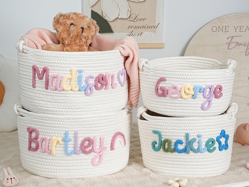 May include: Four white rope baskets with embroidered names: Madison, George, Bartley, and Jackie. The baskets are filled with a teddy bear and a pink blanket. The names are in pastel colors. A rainbow and a star are also embroidered.