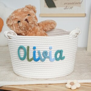 May include: A white rope basket with the name "Olivia" in blue and green yarn. A brown teddy bear sits inside the basket. The basket is on a wooden surface.