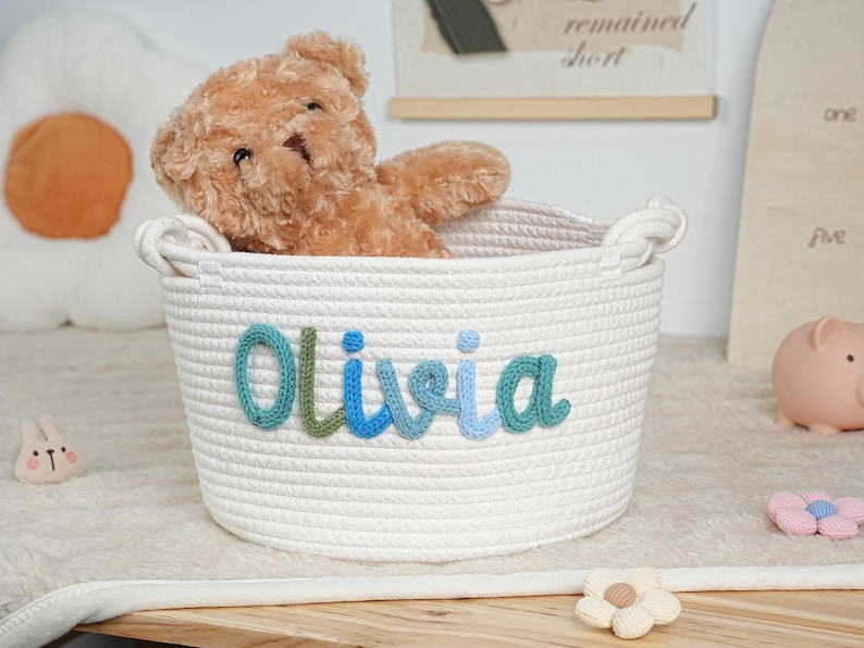 May include: A white rope basket with the name "Olivia" in blue and green yarn. A brown teddy bear sits inside the basket. The basket is on a wooden surface.