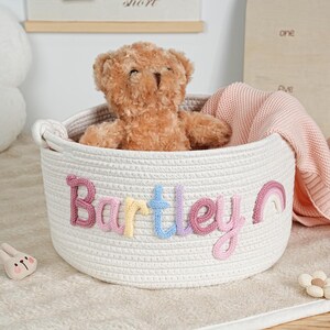May include: A white rope basket with the name "Bartley" in colorful stitched letters. A brown teddy bear and a pink blanket are inside. A small pink piggy bank and a flower-shaped toy are nearby.