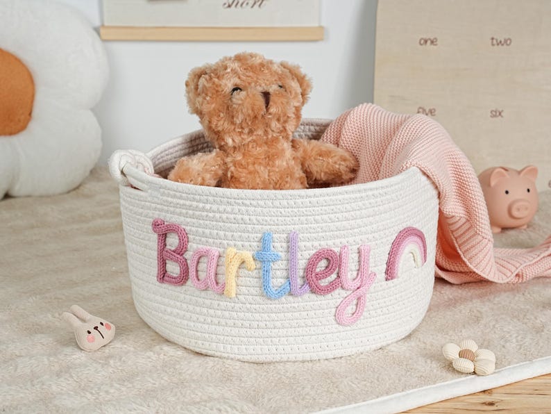 May include: A white rope basket with the name "Bartley" in colorful stitched letters. A brown teddy bear and a pink blanket are inside. A small pink piggy bank and a flower-shaped toy are nearby.