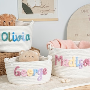 May include: Three white rope baskets with embroidered names: "Olivia", "George", and "Madison". Each basket has a different colored name. The baskets are filled with a teddy bear and a pink blanket. A wooden sign is in the background.