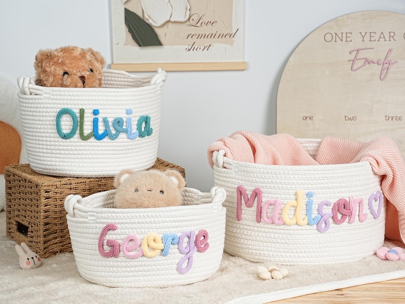 May include: Three white rope baskets with embroidered names: "Olivia", "George", and "Madison". Each basket has a different colored name. The baskets are filled with a teddy bear and a pink blanket. A wooden sign is in the background.