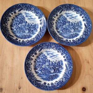 May include: Three round ceramic plates featuring a blue and white transferware pattern. Each plate displays a detailed pastoral landscape scene in the center, surrounded by a decorative border of flowers, leaves, and wheat stalks.