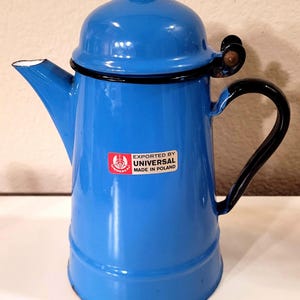 May include: A blue enamel coffee pot with a black handle and trim. The pot has a spout and a lid with a small knob. A sticker on the front reads "EXPORTED BY UNIVERSAL MADE IN POLAND."