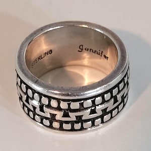 May include: A silver band ring featuring a repeating geometric pattern around the exterior. The interior of the ring is engraved with the word STERLING and the name Jennifer in a cursive script font.