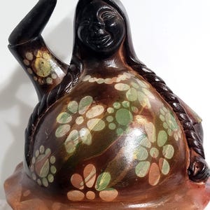 May include: A hand-painted, wooden figurine of a woman carrying a bundle on her head. The figure has dark skin, long braids, and is adorned with floral patterns in green, yellow, and white on a brown background. The base is a darker brown.