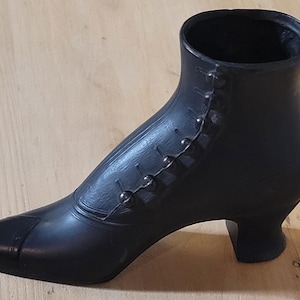 May include: A black decorative boot-shaped object featuring a pointed toe, a curved heel, and a vertical row of small round buttons running along the side. The item has a smooth, matte finish and an open top, resembling a vintage-style ankle boot.