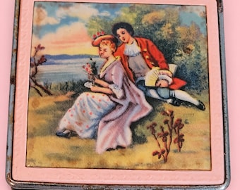 May include: A square metal compact case featuring a light pink border and a central illustration of two figures in period clothing sitting in a landscape with a body of water in the background. The metal edges show signs of wear and aging.