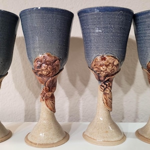 May include: Four handmade ceramic goblets. The cups are a muted blue with a textured finish. The stems are beige with a detailed floral and leaf design in brown. The goblets are displayed on a white surface.