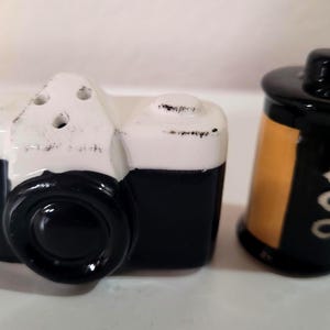 May include: A ceramic salt and pepper shaker set shaped like a vintage camera and a film canister. The camera is black and white with a large lens. The film canister is black with a yellow label and the number 400.