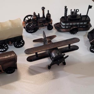 May include: A collection of miniature antique vehicles, including a covered wagon, stagecoach, steam engine, boat, and biplane. The models are made of a dark metallic material and have intricate details.