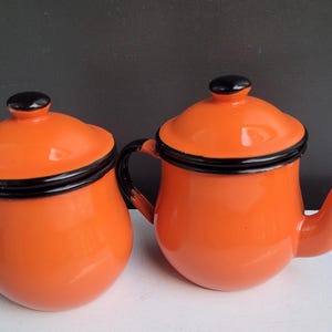 May include: Two orange enamelware kitchen items: a small covered jar and a teapot. Both have black trim and lids. The teapot has a curved spout and a black handle. The items are set against a dark background.