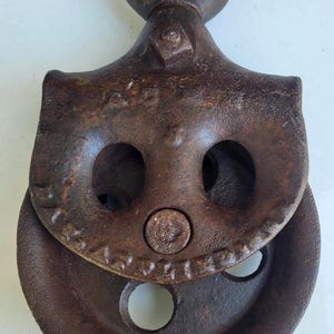 Puede incluir: Polea de metal oxidado y envejecido, con un gran lazo superior circular. La polea tiene una rueda y dos aberturas circulares. El metal muestra signos de edad y uso. Esta polea antigua probablemente se utiliza para levantar o tirar.