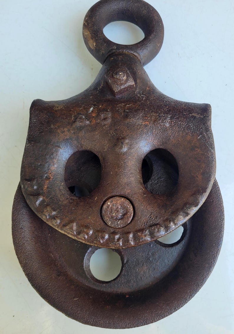 Puede incluir: Polea de metal oxidado y envejecido, con un gran lazo superior circular. La polea tiene una rueda y dos aberturas circulares. El metal muestra signos de edad y uso. Esta polea antigua probablemente se utiliza para levantar o tirar.