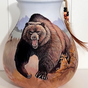 May include: A gourd vessel featuring a hand-painted illustration of a brown grizzly bear with an open mouth and bared teeth. The scene includes a mountain landscape and autumn foliage. A leather cord with multi-colored beads and a feather is attached.