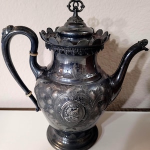 May include: Antique silver-plated teapot with intricate floral and medallion detailing. The teapot features a curved handle, a decorative spout, and a lid with a finial. The design includes engraved patterns and a detailed base, showcasing a classic, ornate style.