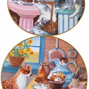 May include: Two round decorative plates featuring cat illustrations. The top plate shows kittens in a bathroom with a pink sink. The bottom plate depicts orange cats in a kitchen with a blue pitcher, a bowl of milk, and sunflowers in a basket.